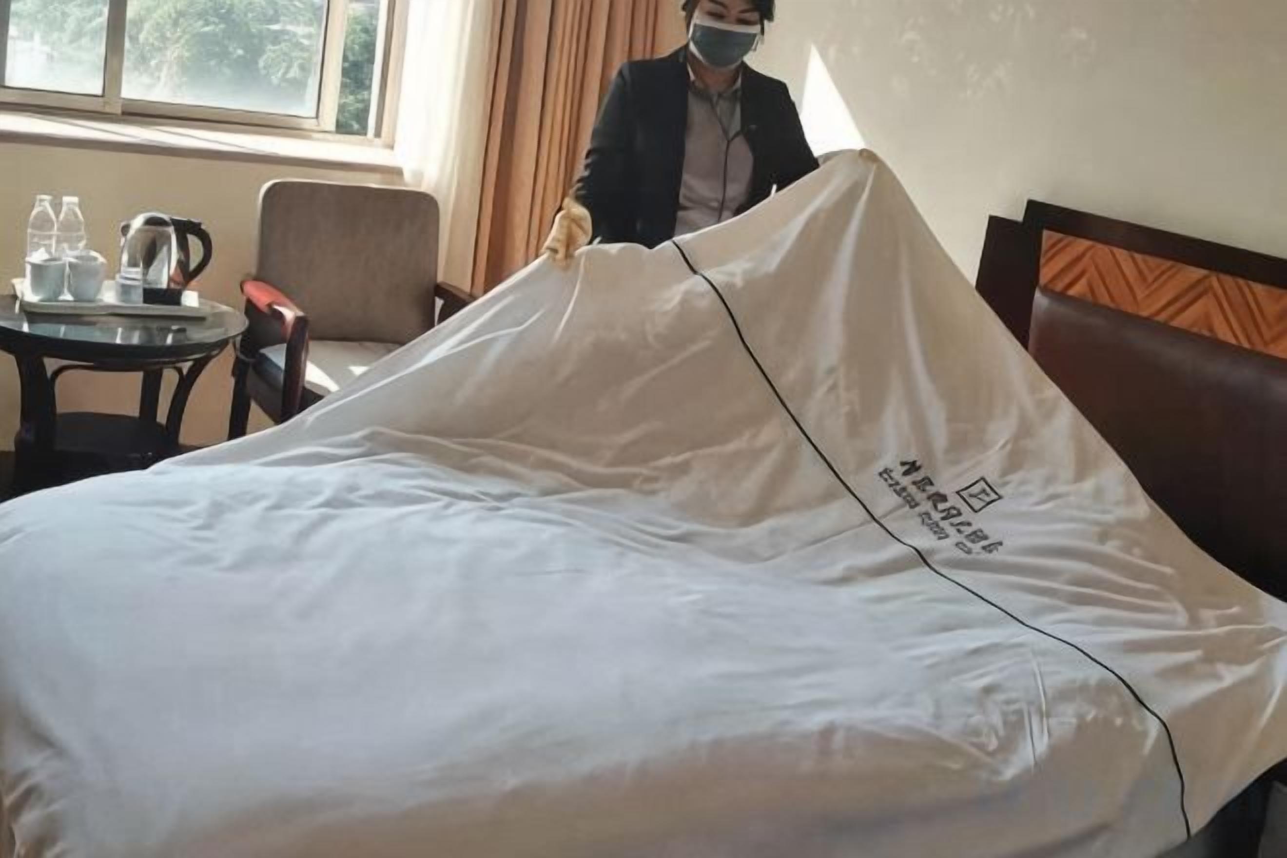 Ein Hotelzimmer mit einem Bett, das mit einem weißen Laken bezogen ist, einem Nachttisch mit Wasserkocher und Tassen sowie einem Sessel.
