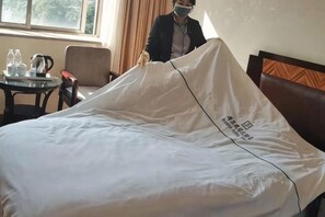 A hotel room with a bed covered in a white sheet, a bedside table with a kettle and cups, and a chair.