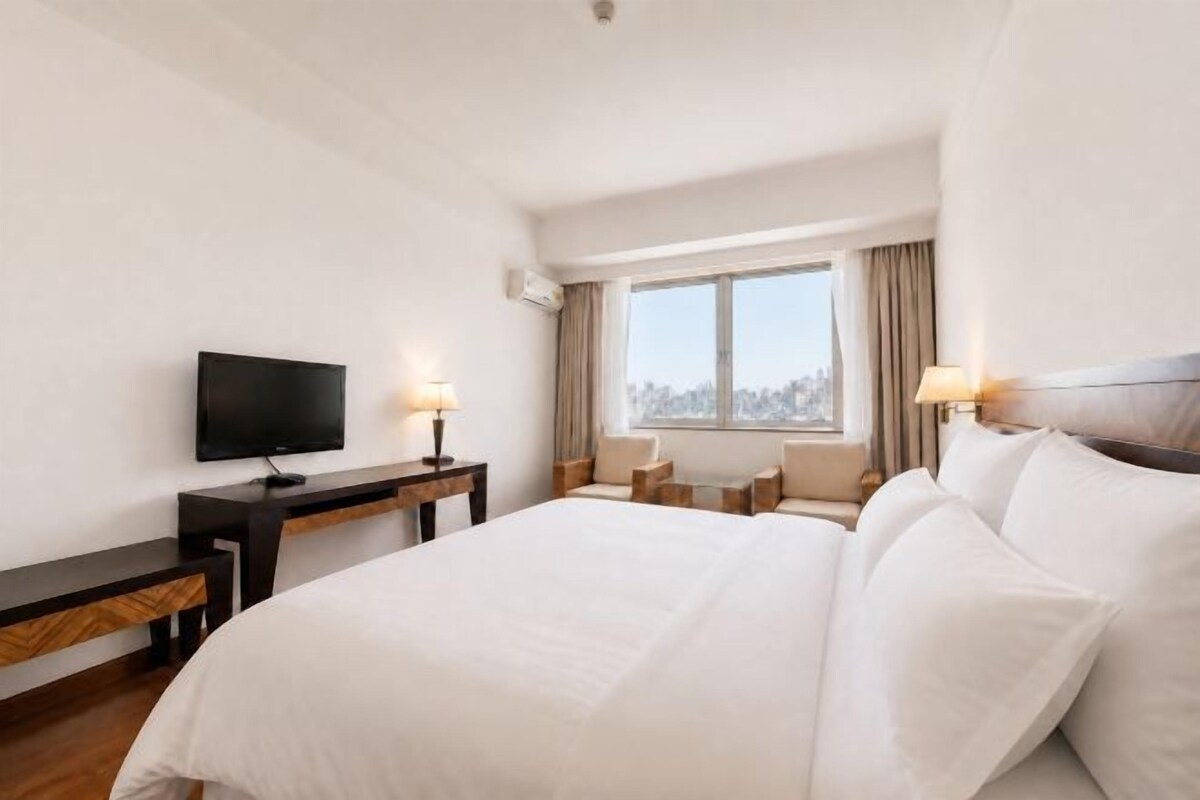 Habitació d'hotel amb un llit gran, un televisor de pantalla plana, un escriptori amb una làmpada i una finestra amb vista a la ciutat.