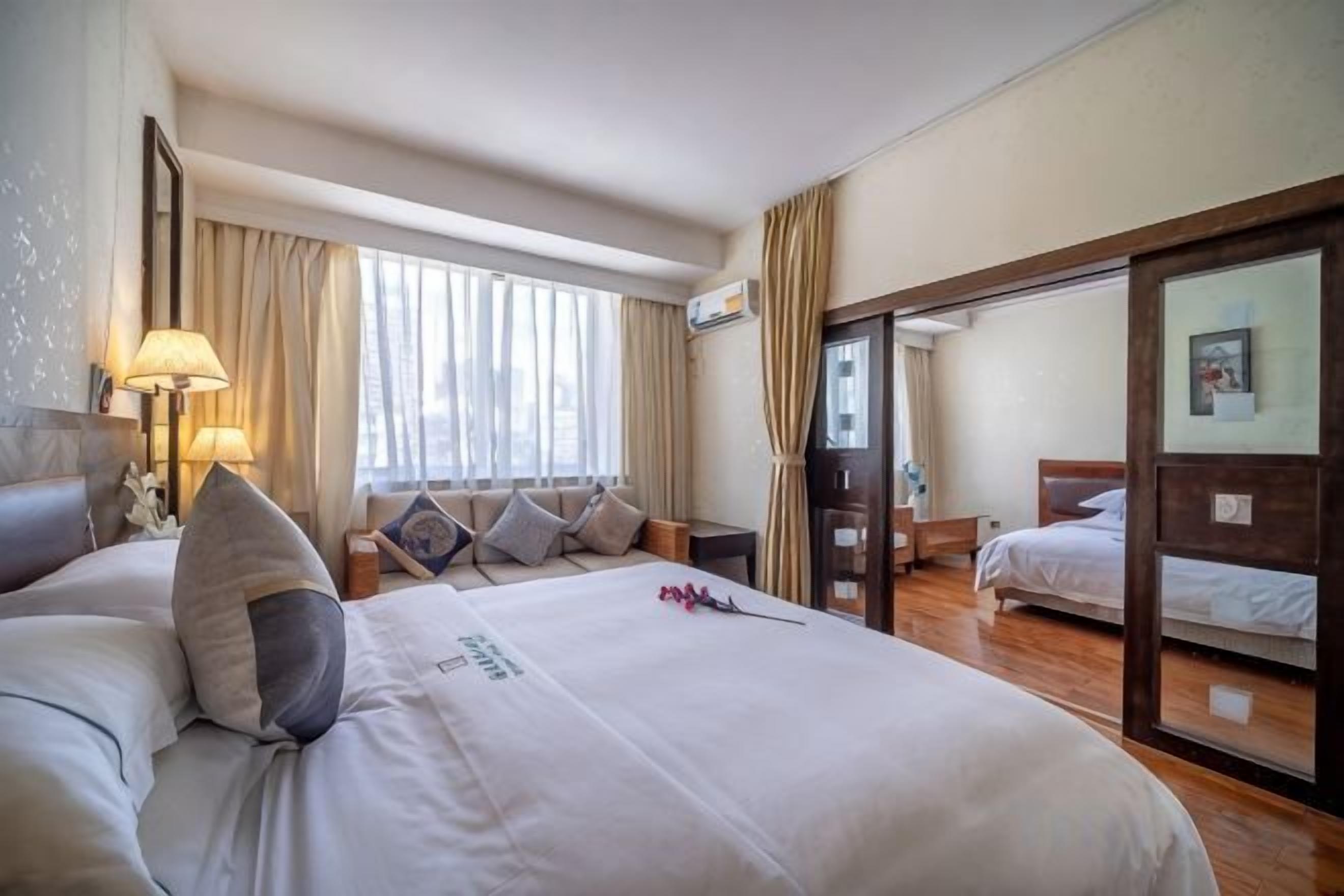 A hotel room with a large bed, a desk, a chair, a window with curtains, and a door leading to another room.