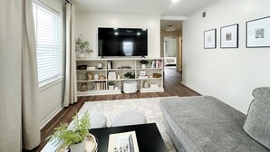A living room with a grey sofa, a black coffee table, and a TV mounted on a white wall. There are shelves with decorative items and a window with blinds.