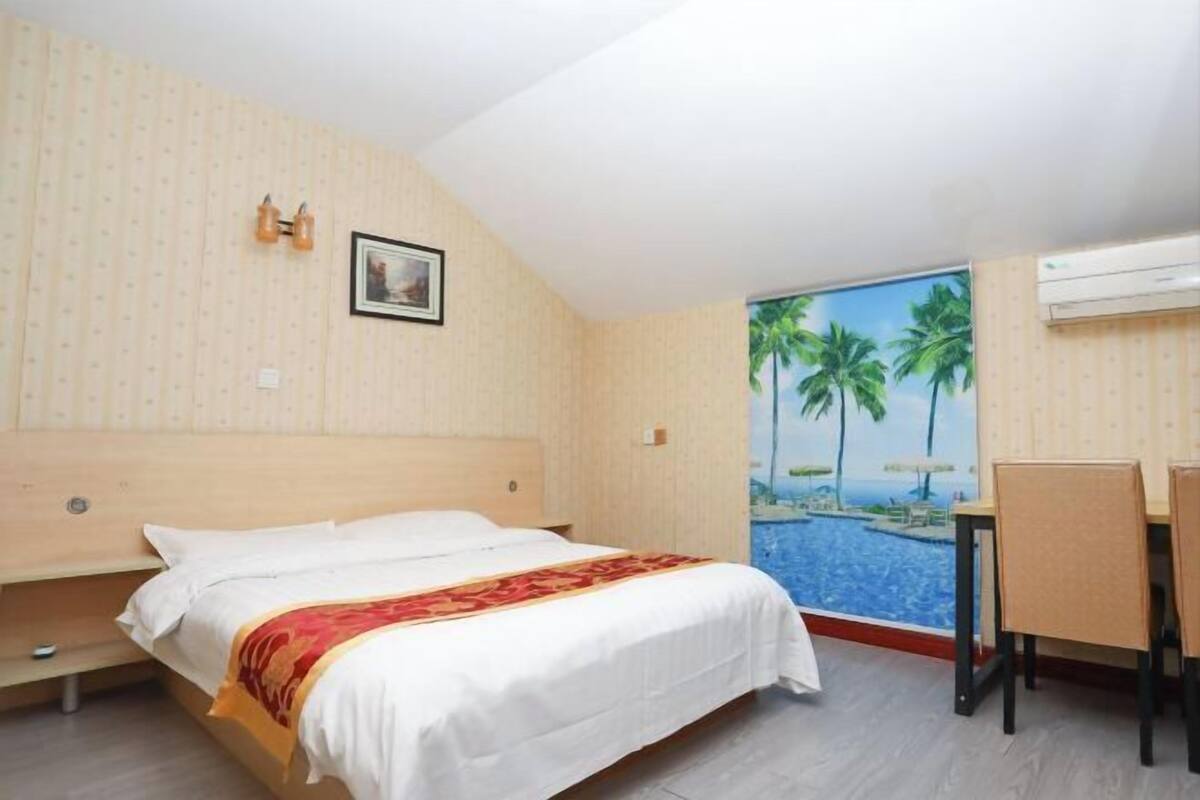 A hotel room with a bed, a desk with chairs, a painting on the wall, and an air conditioning unit.