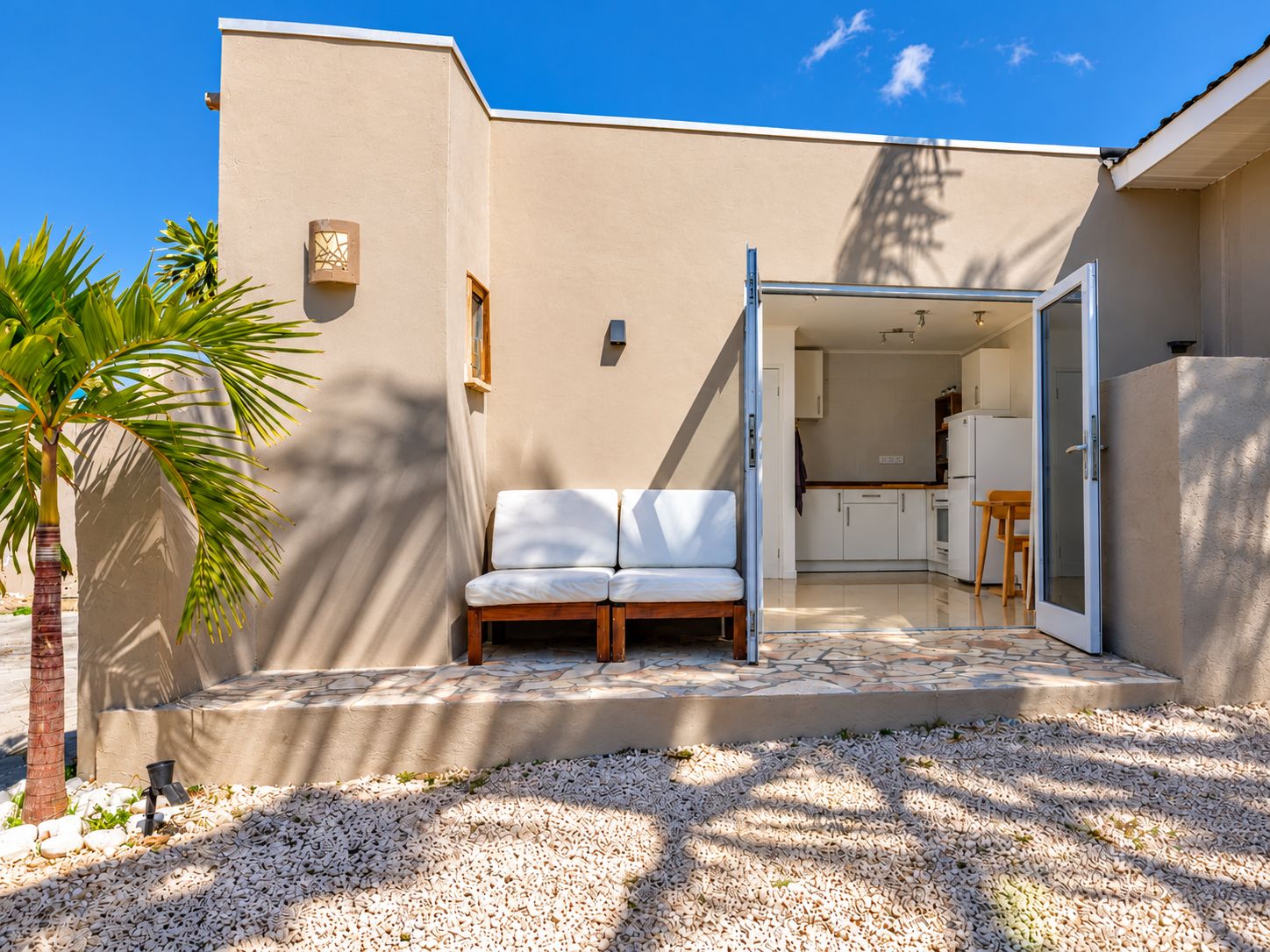 Et moderne udendørs opholdsområde med en hvid sofa, et palmetræ og en grussti, der fører til en glasdør.