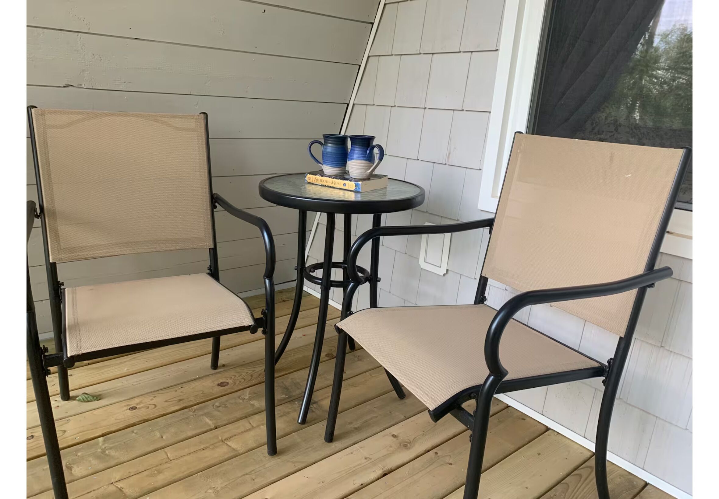 Un pequeño espacio al aire libre con dos sillas y una mesa, mostrando un libro y dos tazas azules.