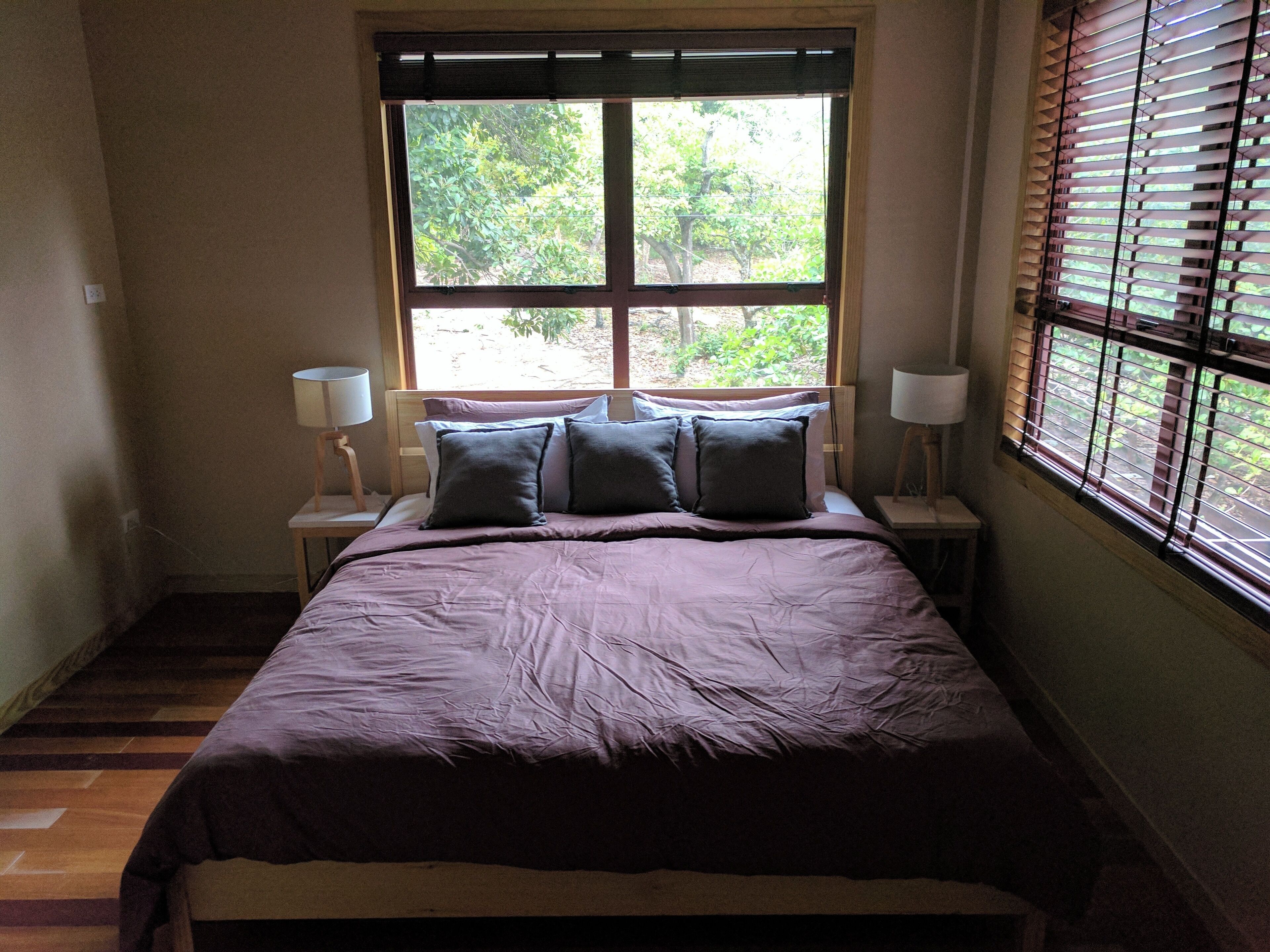 Un dormitorio con un ventanal grande, una cama con colcha morada, dos mesitas de noche con lámparas y vistas al exterior con vegetación.