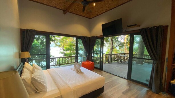 A bedroom with a large bed, a ceiling fan, a TV, and a balcony with a view of trees and a body of water. - Cashewnut Cove Bungalow - Beach Front (Ko Lipe)