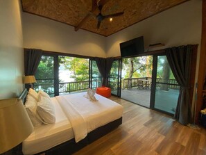 Ein Schlafzimmer mit einem großen Bett, einem Deckenventilator, einem Fernseher und einem Balkon mit Blick auf Bäume und ein Gewässer.