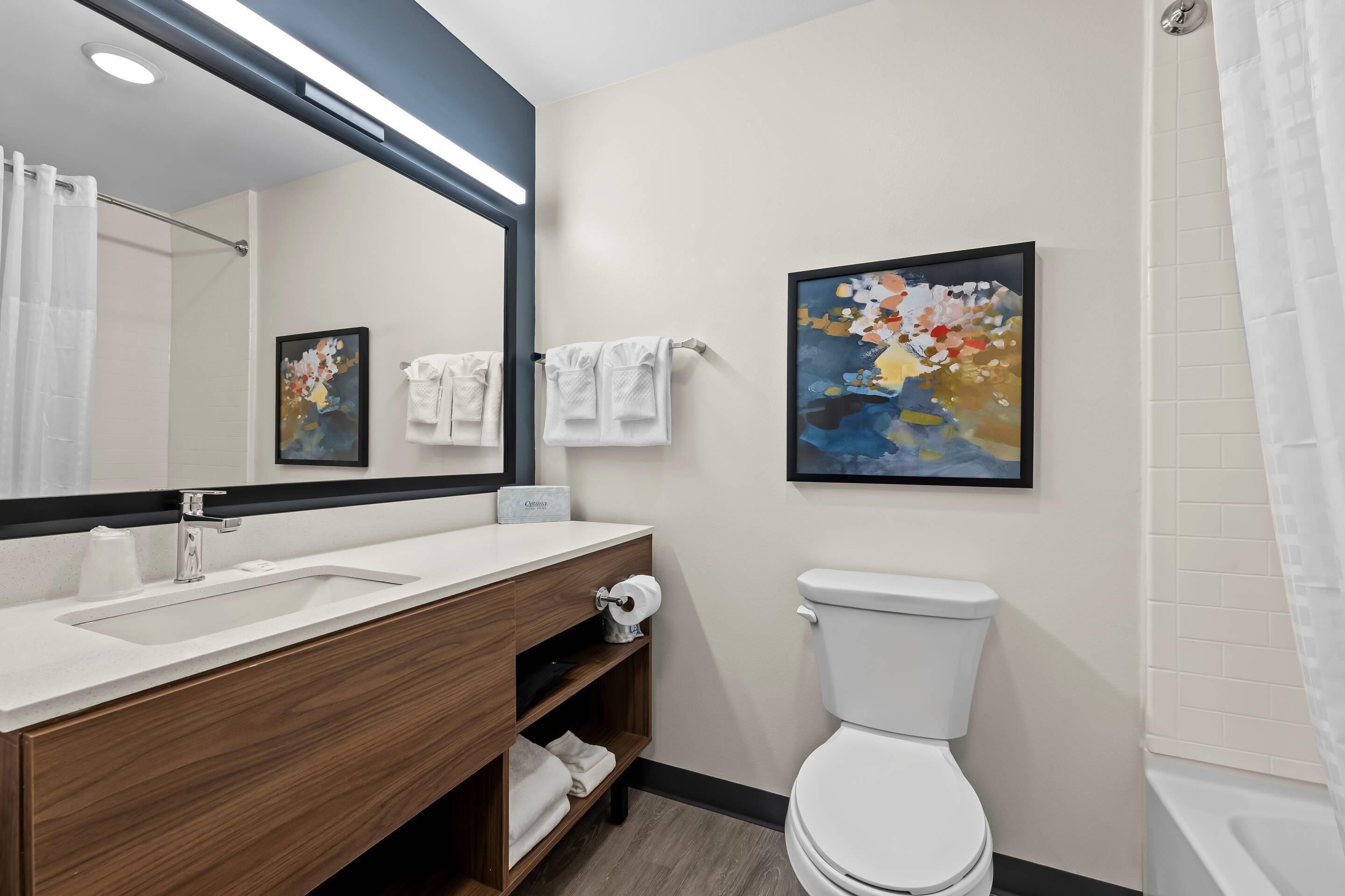 A modern bathroom with a wooden vanity, white sink, and a large mirror. There is a toilet and a shower area with a white curtain.