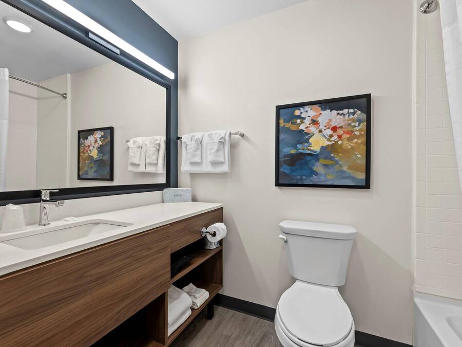 A modern bathroom with a wooden vanity, white sink, and a large mirror. There is a toilet and a shower area with a white curtain.