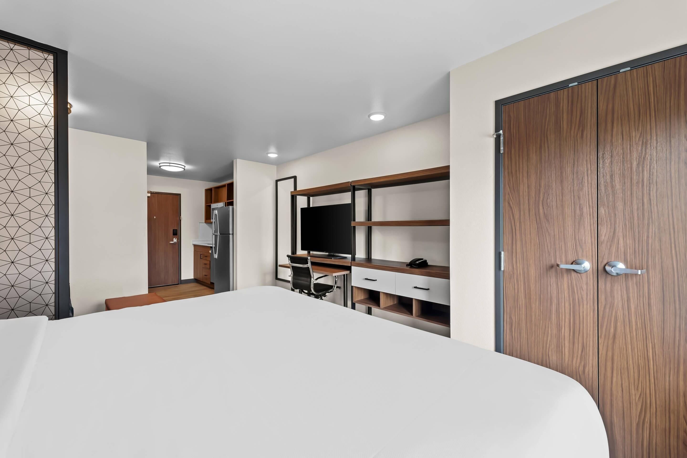 A modern hotel room with a large bed, a desk with a chair, a wardrobe, and a kitchenette.