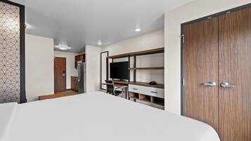 A modern hotel room with a large bed, a desk with a chair, a wardrobe, and a kitchenette.