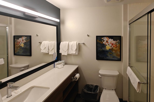 Un baño moderno con un espejo grande, lavamanos blanco y dos inodoros. Hay toallas colgadas en la pared y un bote de basura.
