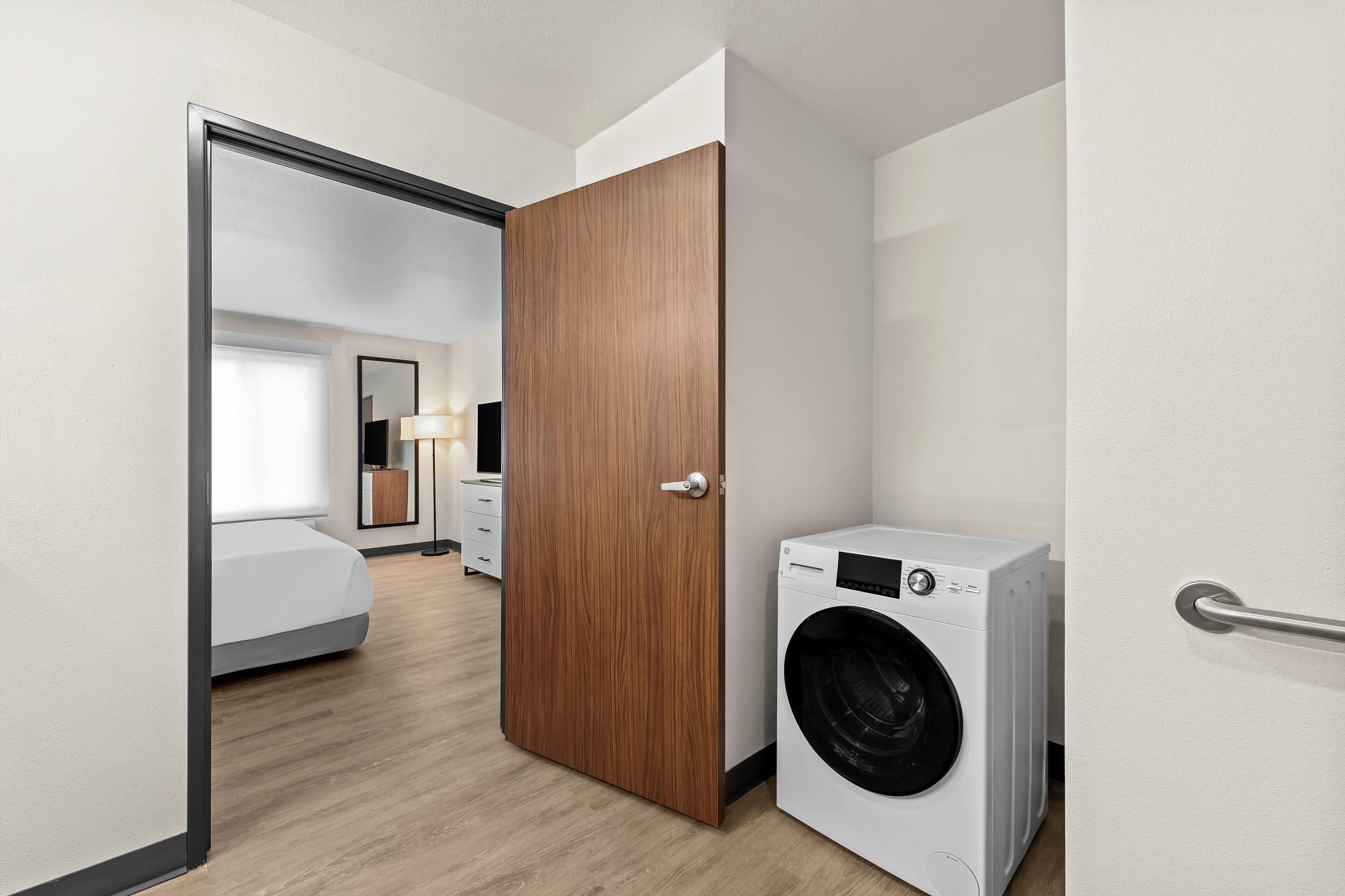 A hotel room with a wooden door, a washing machine, a bed, and a bedside table.