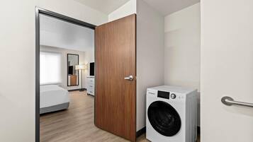 A hotel room with a wooden door, a washing machine, a bed, and a bedside table.