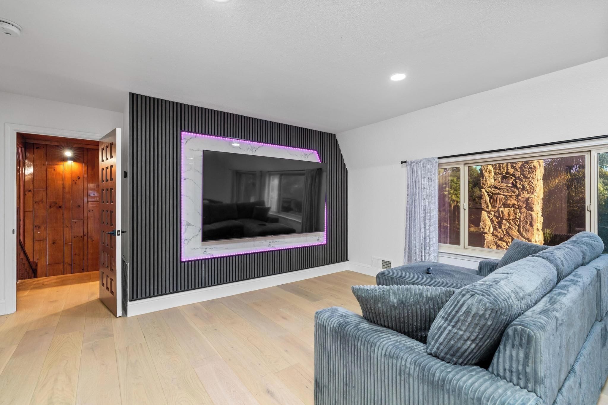 A modern living room with a large flat-screen TV mounted on a black wall, a grey sofa, and a window with a view of a rocky landscape.
