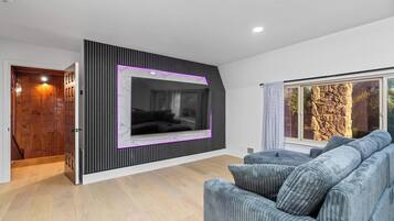 A modern living room with a large flat-screen TV mounted on a black wall, a grey sofa, and a window with a view of a rocky landscape.