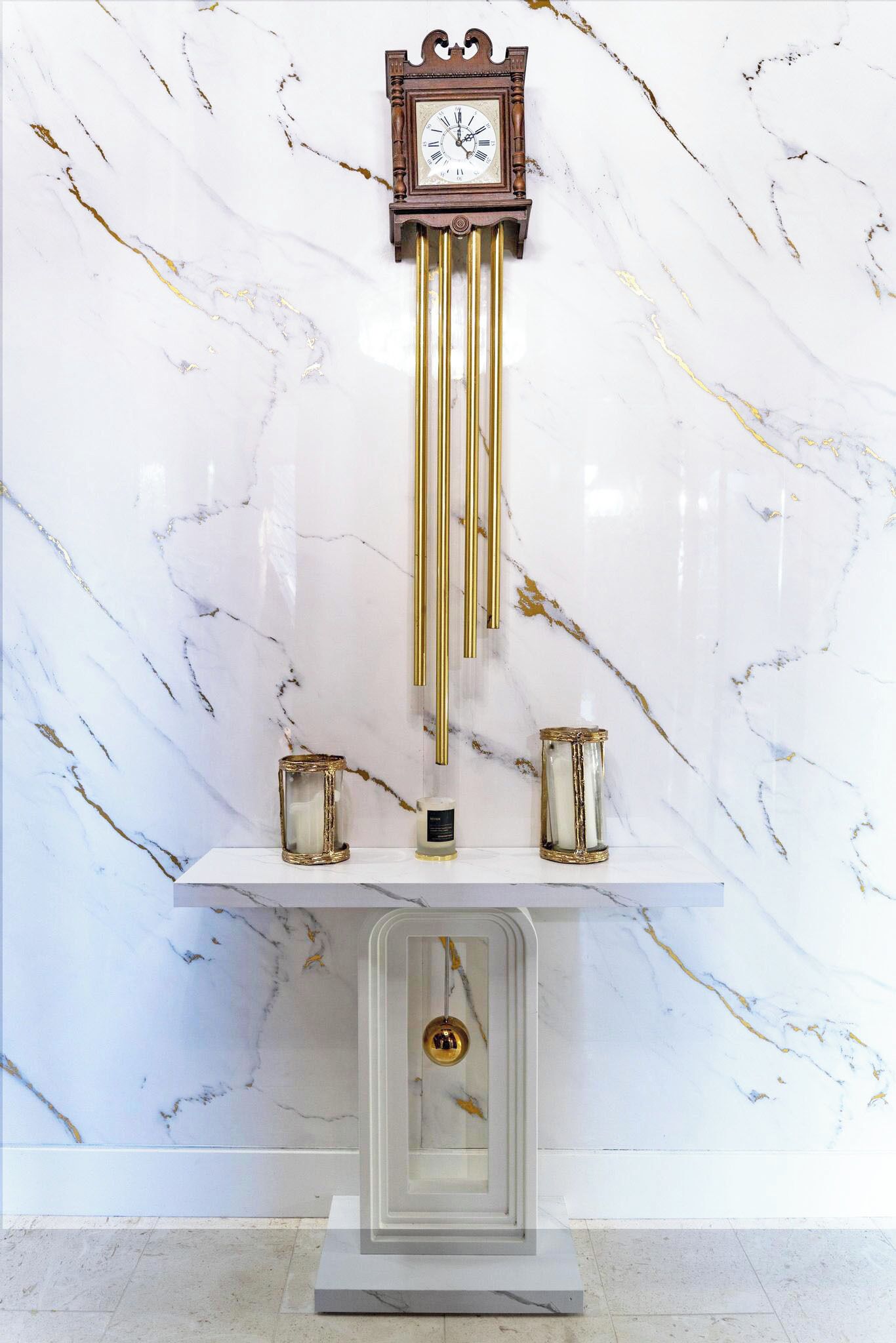 Pared de mármol con un reloj y dos candelabros sobre un pedestal.