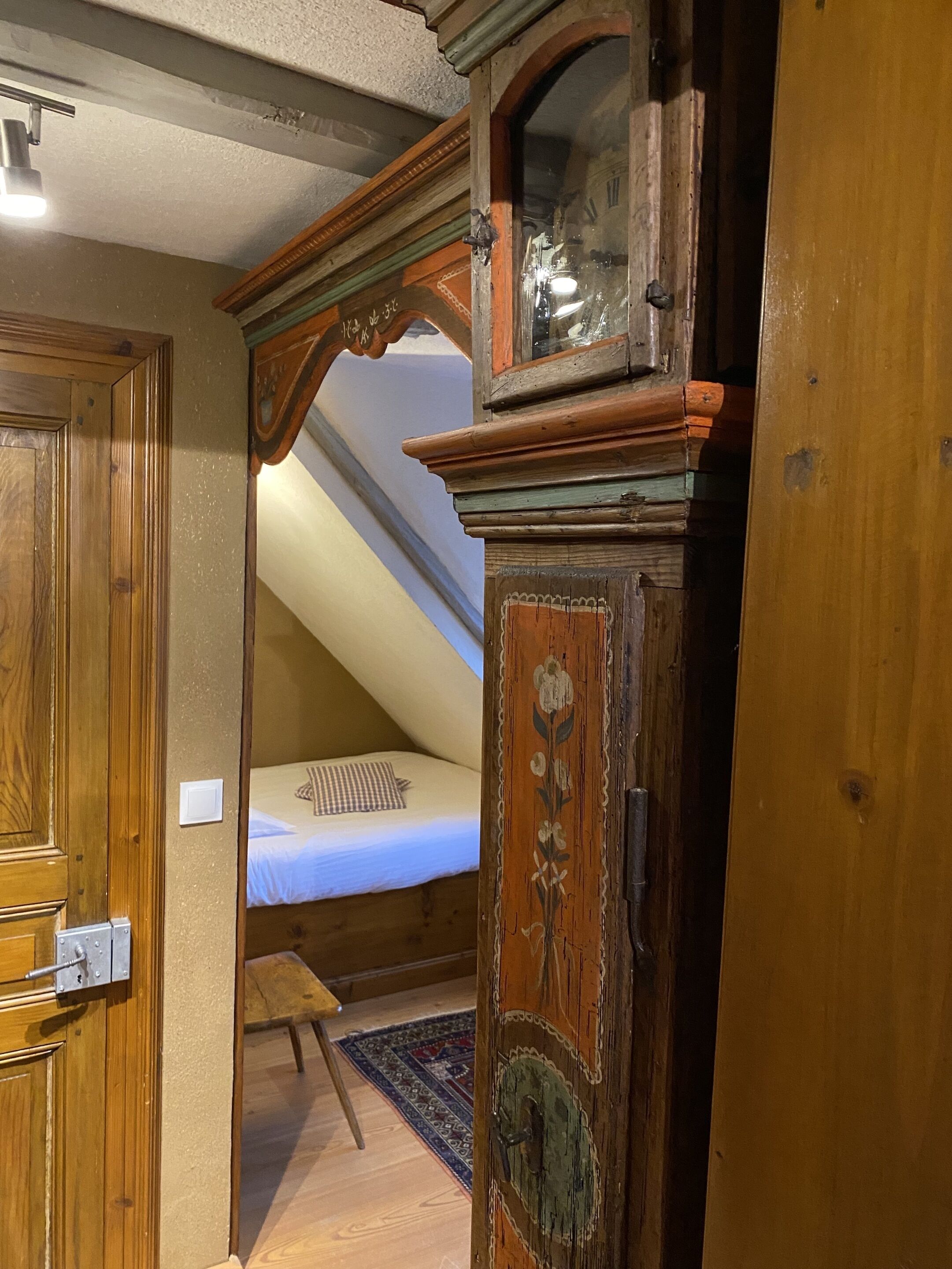 Une pièce avec une porte en bois, un lit recouvert d’une courtepointe à carreaux, un petit tabouret et un tapis à motifs.