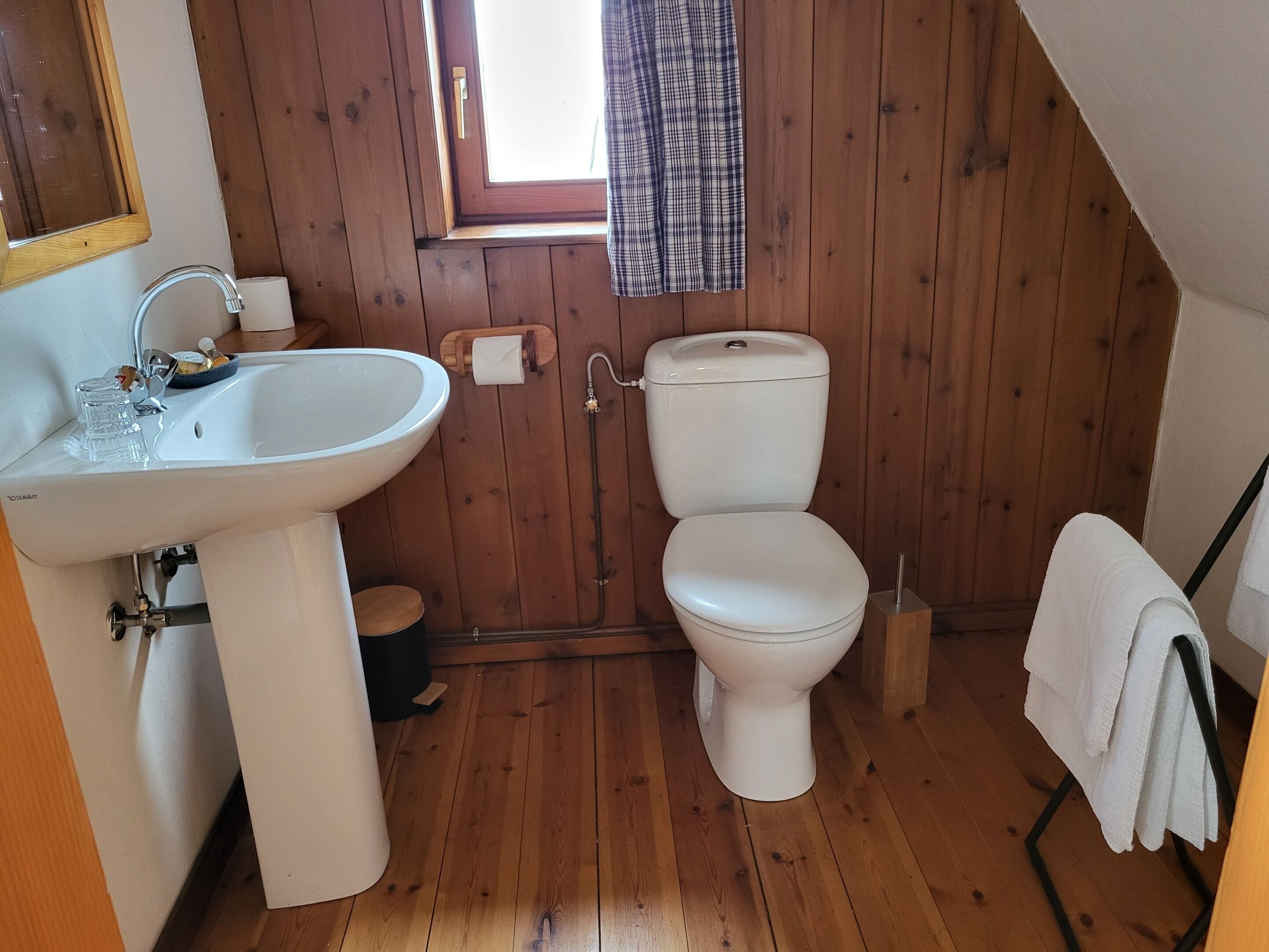 A small, wooden-paneled bathroom with a white sink, toilet, and a window with a plaid curtain.