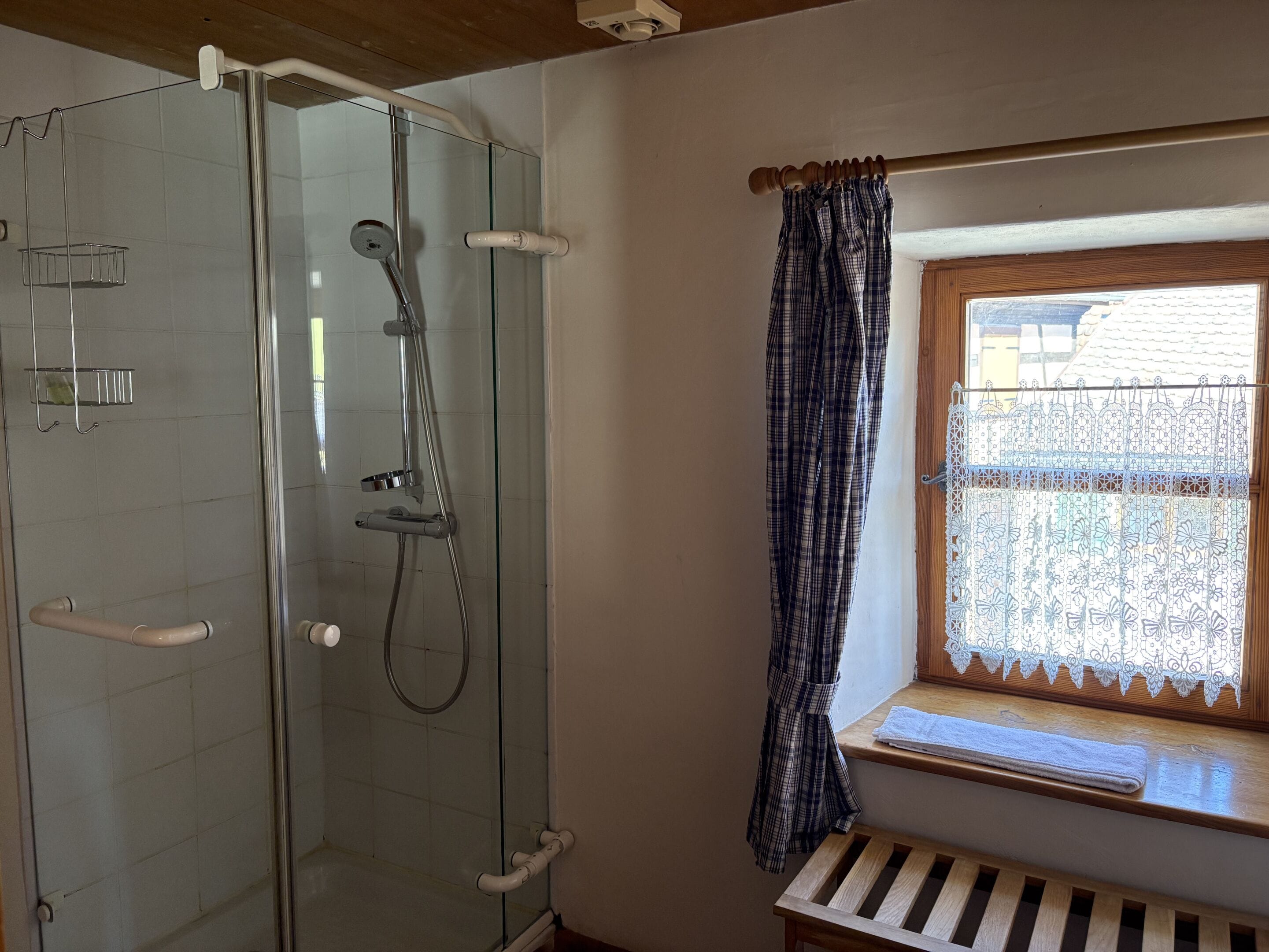 A shower with a glass door, a window with a lace curtain, and a wooden bench.