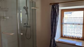 A shower with a glass door, a window with a lace curtain, and a wooden bench.