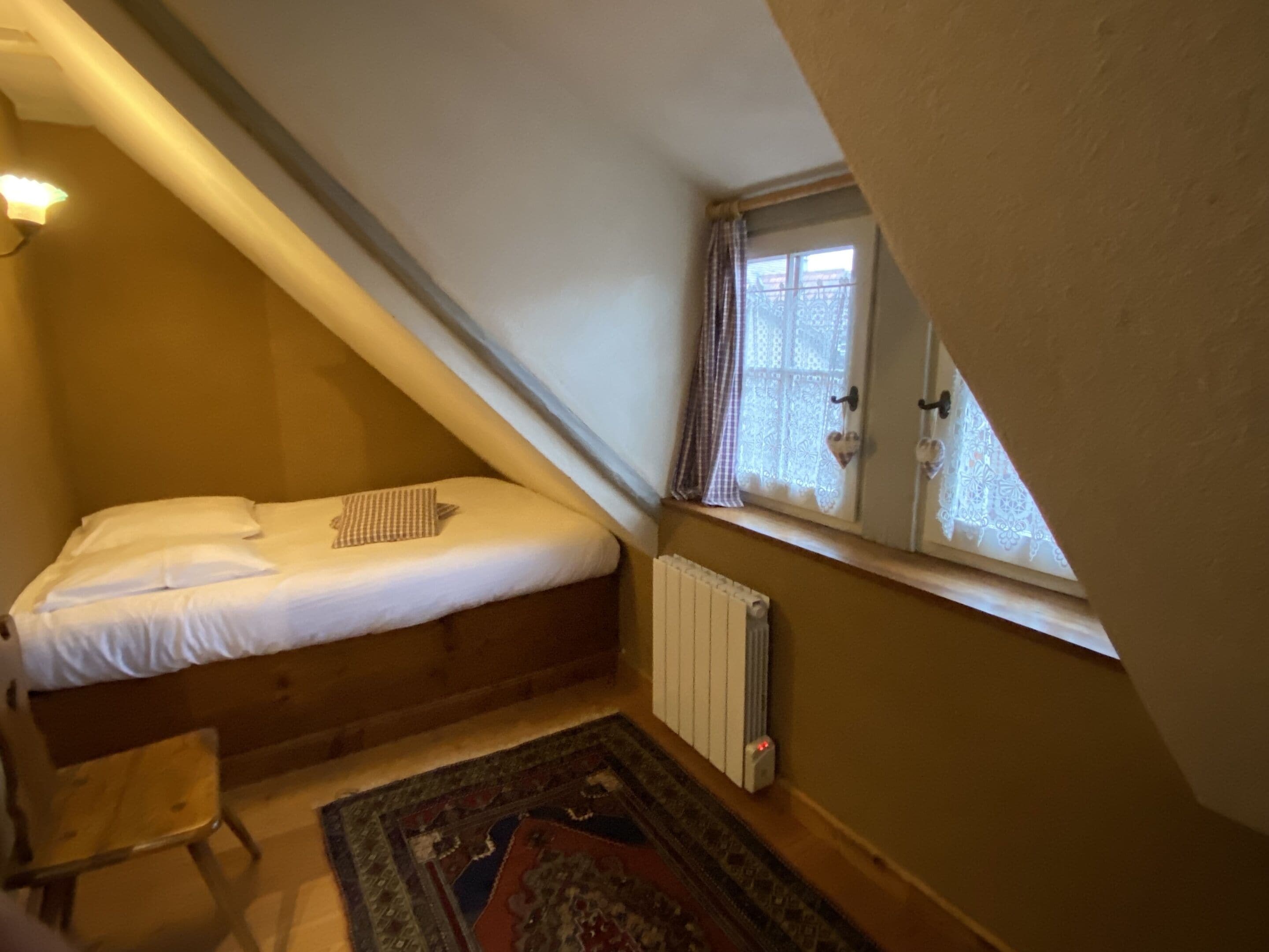 A small, sloped bedroom with a single bed, a radiator, a window with curtains, and a patterned rug.