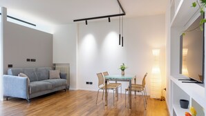 A modern living room with a grey sofa, wooden flooring, a dining table with chairs, a hanging light fixture, and a TV on a white shelf. - A terrace over St. Peter's (Roma)