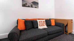 Un sofá gris oscuro con cojines naranja y blancos, una mesita de madera y un cuadro en la pared que muestra una escena costera.