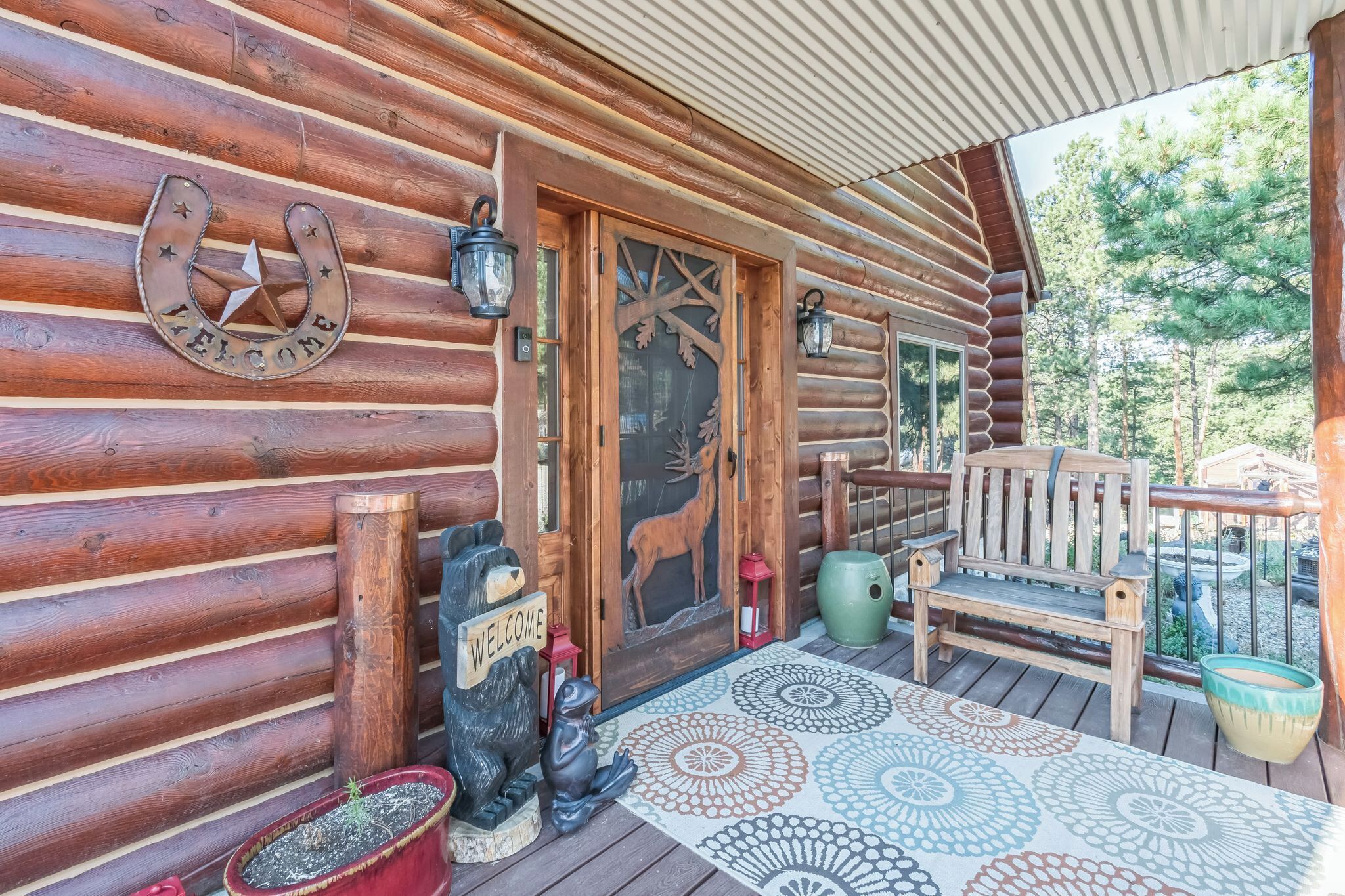 A wooden cabin with a welcome sign, a bear statue, a bench, and a patterned rug on the porch.