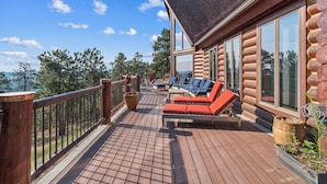 A wooden deck with outdoor seating, large windows, and a view of the surrounding forest. - Luxe Log Home+Views+Hot Tub+Fire Pit+Open Air Game Room + Red Rocks (Bailey)
