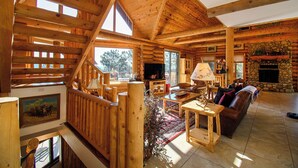 A log cabin interior with a wooden staircase, a stone fireplace, and a large window. - Luxe Log Home+Views+Hot Tub+Fire Pit+Open Air Game Room + Red Rocks (Bailey)