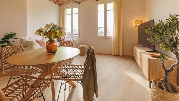 Un salón acogedor con una mesa redonda de madera, sillas de mimbre, una planta grande y un televisor sobre un mueble de madera.