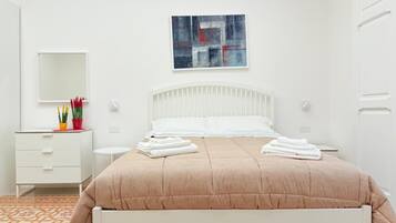 A neatly arranged bedroom with a white bed, a bedside table, a nightstand, a small desk with a chair, and a wall-mounted mirror.