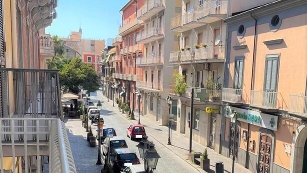 A narrow cobblestone street lined with multi-story buildings, parked cars, and a pedestrian walkway.