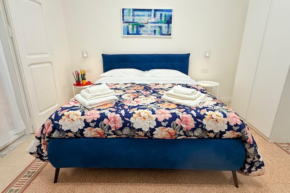 A neatly made bed with a floral duvet, a blue headboard, and a small bedside table with a lamp.