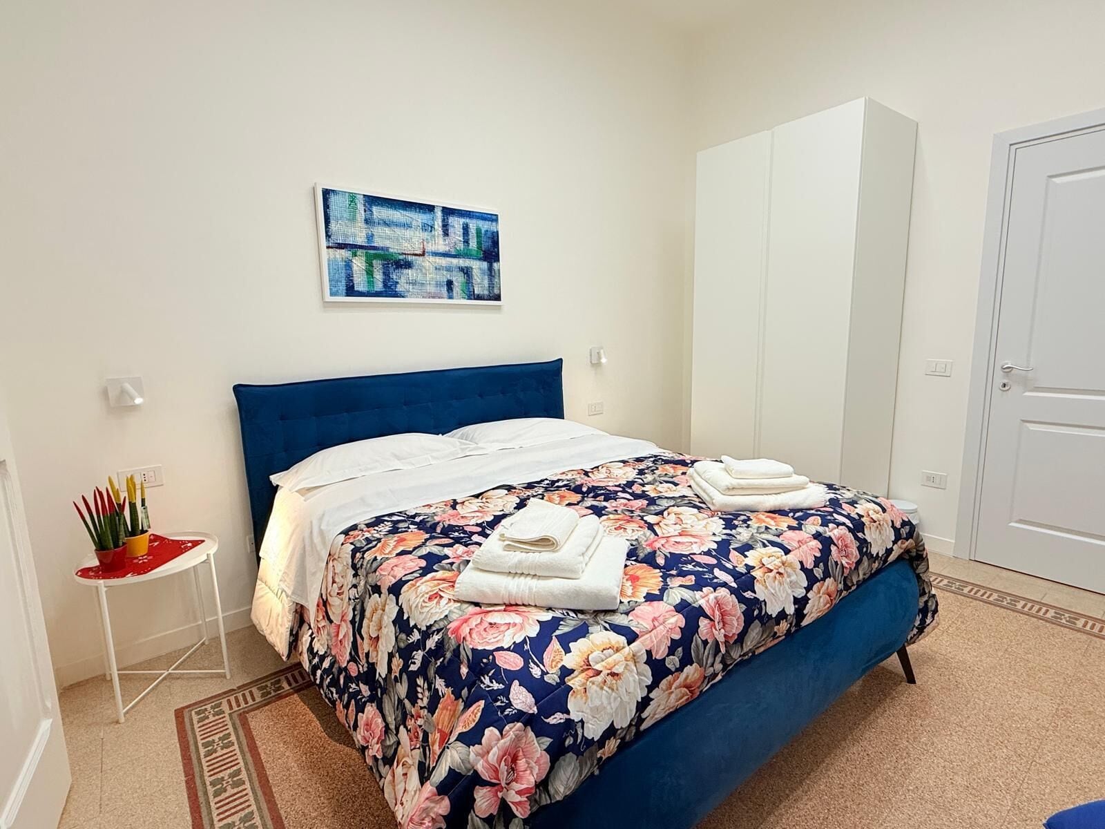 Un dormitorio con una cama, una colcha estampada, un cabecero azul, un cuadro en la pared y un armario blanco.