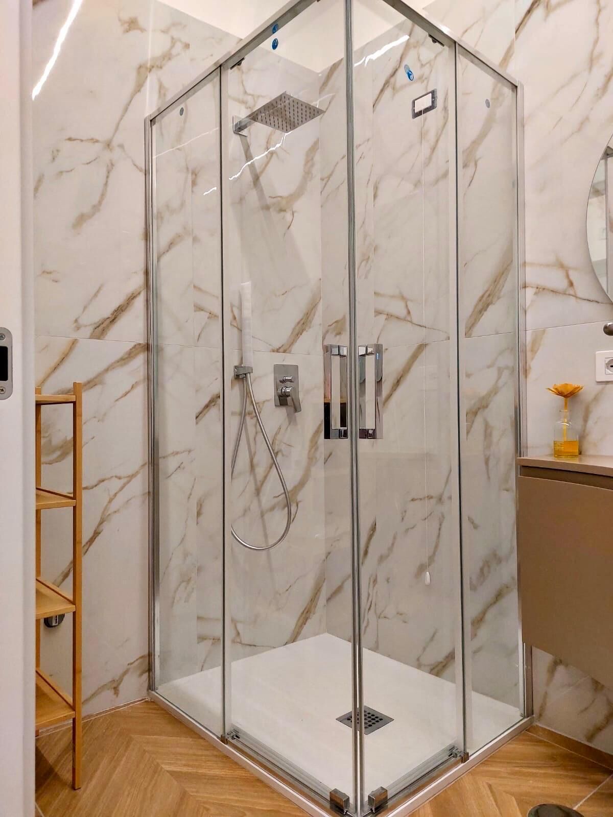 Una ducha con cerramiento de vidrio, pared de mármol, estante de madera y espejo.