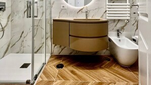 A modern bathroom with a glass shower enclosure, a marble-tiled wall, a wooden floor, a round mirror, a sink with a vanity, and a bidet.