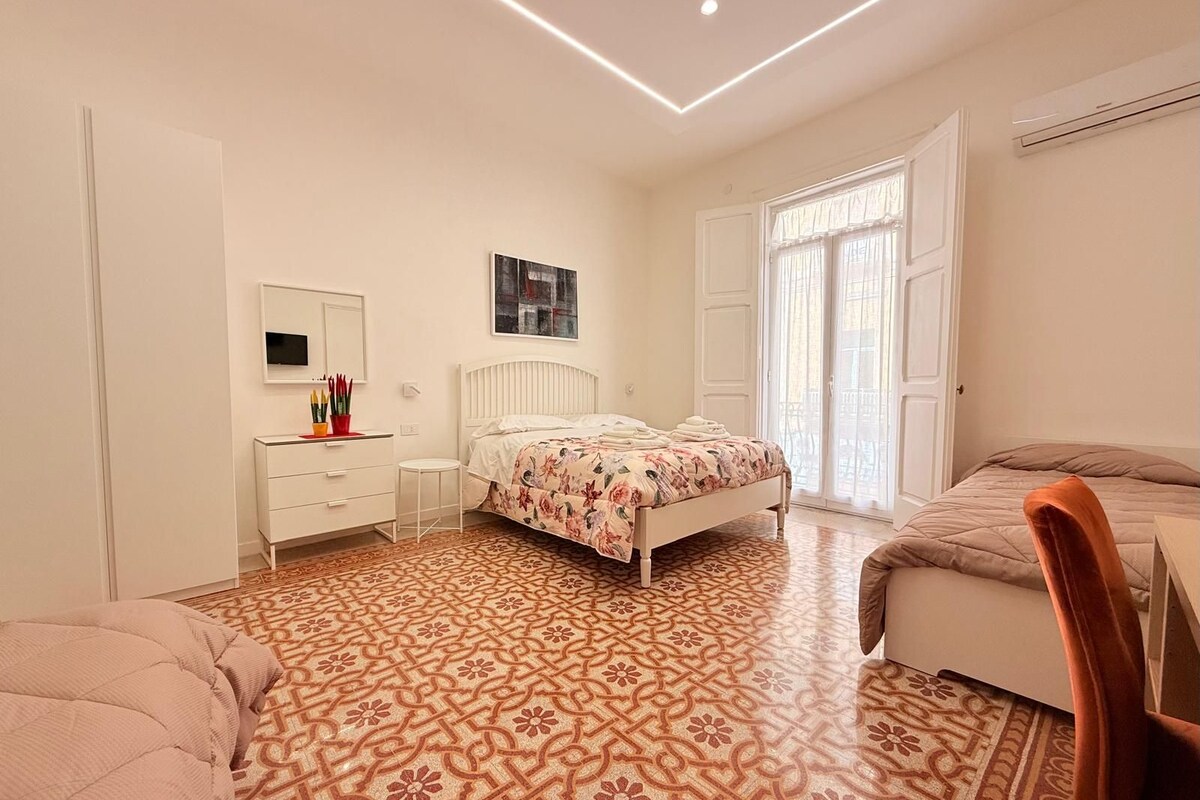 Un dormitorio con piso estampado, una cama con colcha floral, un tocador blanco, una silla y una ventana con cortinas.