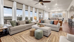 A spacious living room with a large sectional sofa, a ceiling fan, and large windows offering a view of the outdoors. - Pool, Sandy Beach & Amazing Lake Sunsets, Sleeps 16, Family Retreat + Game Room (Kingsland)