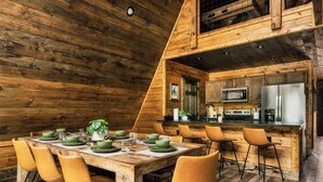 A wooden interior with a dining table set for six, surrounded by chairs, and a kitchen area with modern appliances.