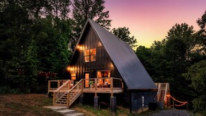 A wooden cabin with a steeply pitched roof, a wooden deck with stairs, and a porch with chairs.