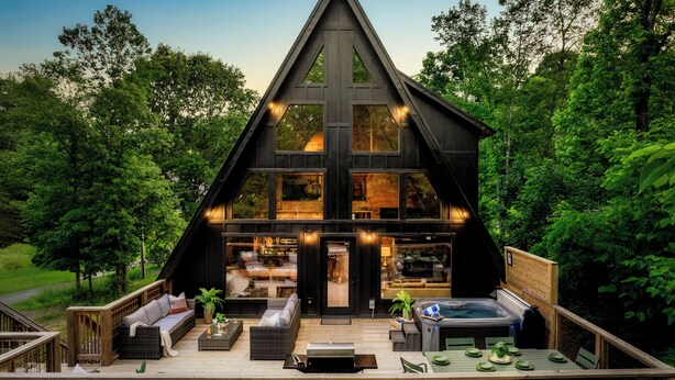 A modern, triangular wooden cabin with a deck, furnished outdoor seating, and a hot tub.