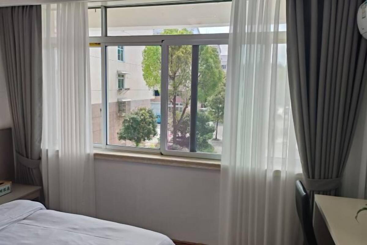 Ein Hotelzimmer mit einem Bett, einem Fenster mit Vorhängen und Blick auf Gebäude und Bäume.