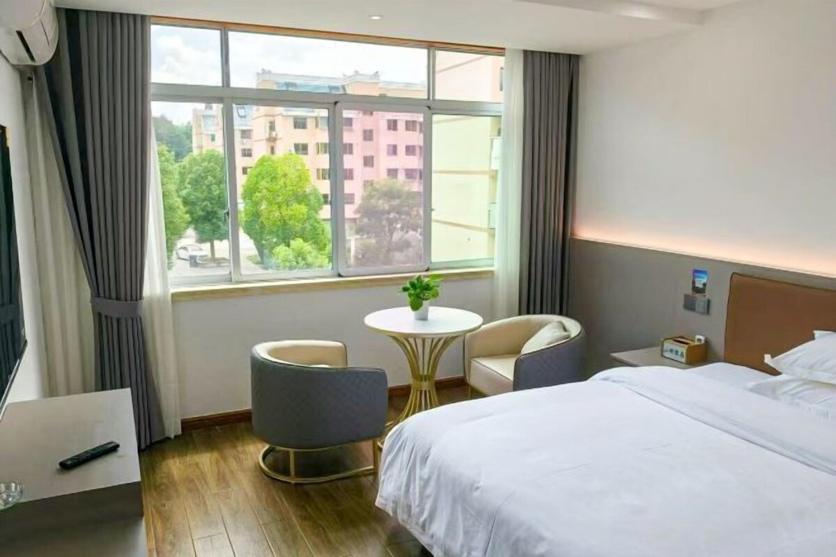 Une chambre d’hôtel comprenant un lit, deux fauteuils, une petite table avec une plante, une table de chevet avec une lampe et une fenêtre donnant sur des bâtiments et des arbres.