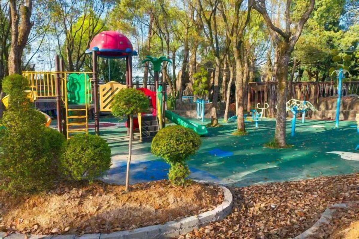 Ein Spielplatz mit verschiedenen Spielgeräten, darunter eine Rutsche und Kletterbereiche, umgeben von Bäumen und einer gepflegten Grünfläche mit geschnittenen Büschen.