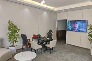 A modern room with a flat-screen TV mounted on the wall, a gaming chair, a dining table with chairs, and a potted plant.