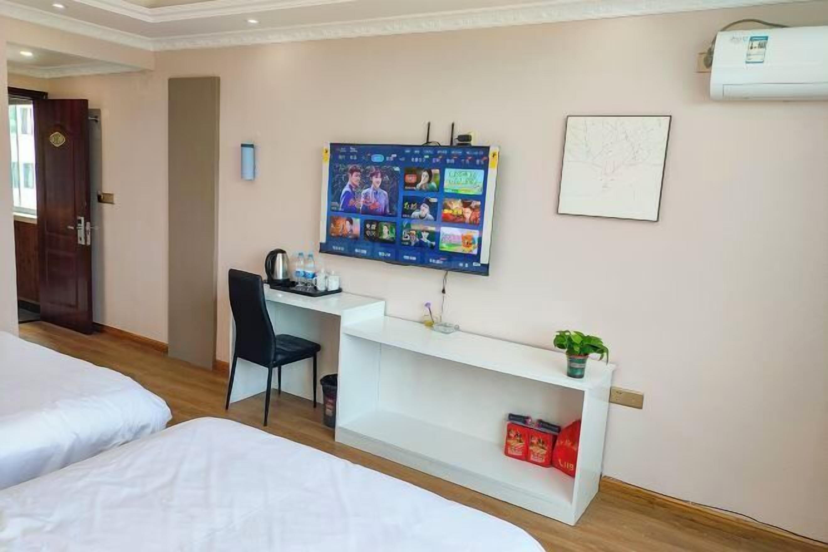 A hotel room with a bed, a desk with a computer, a chair, and a shelf with a plant and some items.