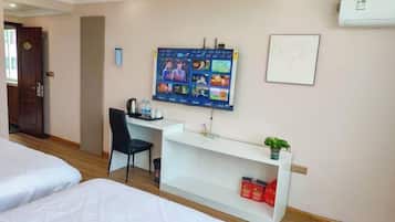 A hotel room with a bed, a desk with a computer, a chair, and a shelf with a plant and some items.