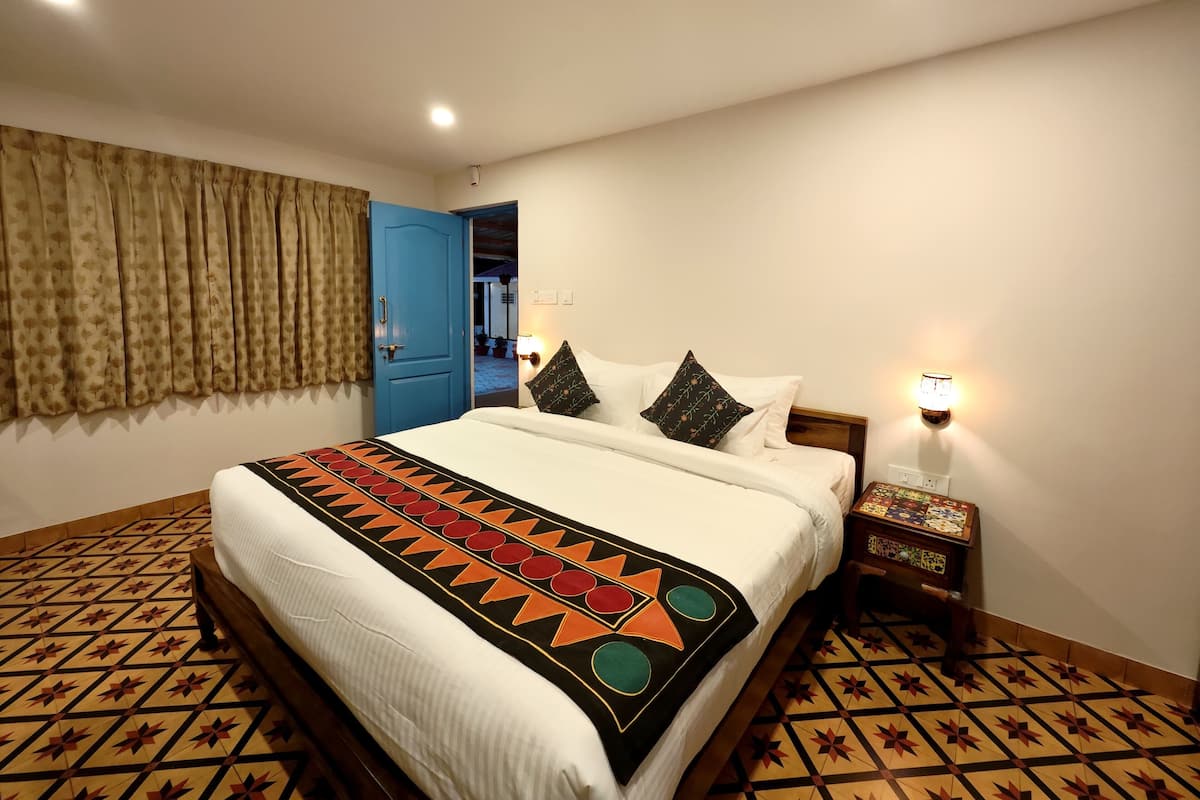 A hotel room with a bed, patterned floor, a blue door, and a nightstand with a lamp.